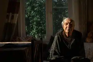 Une personne âgée assise près d'une fenêtre ouverte avec un ventilateur et une bouteille d'eau à proximité