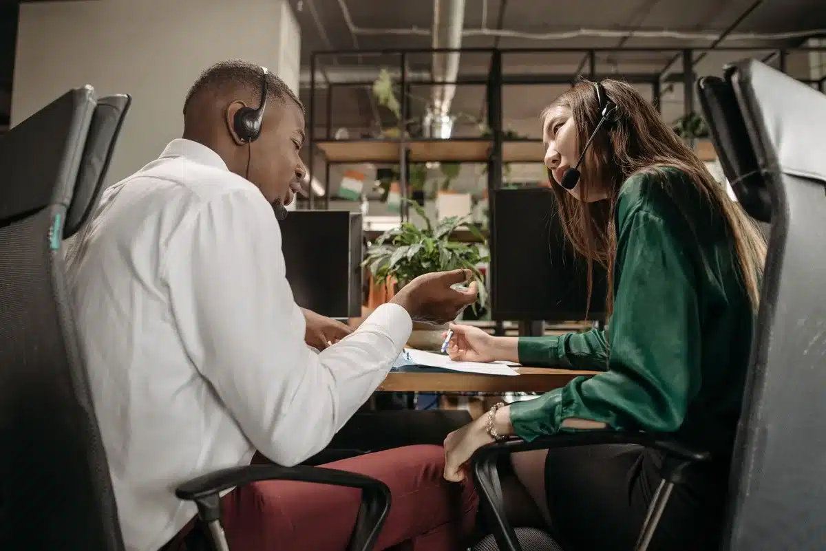 Deux collègues de bureau discutent discrètement à leurs postes de travail dans un environnement professionnel.