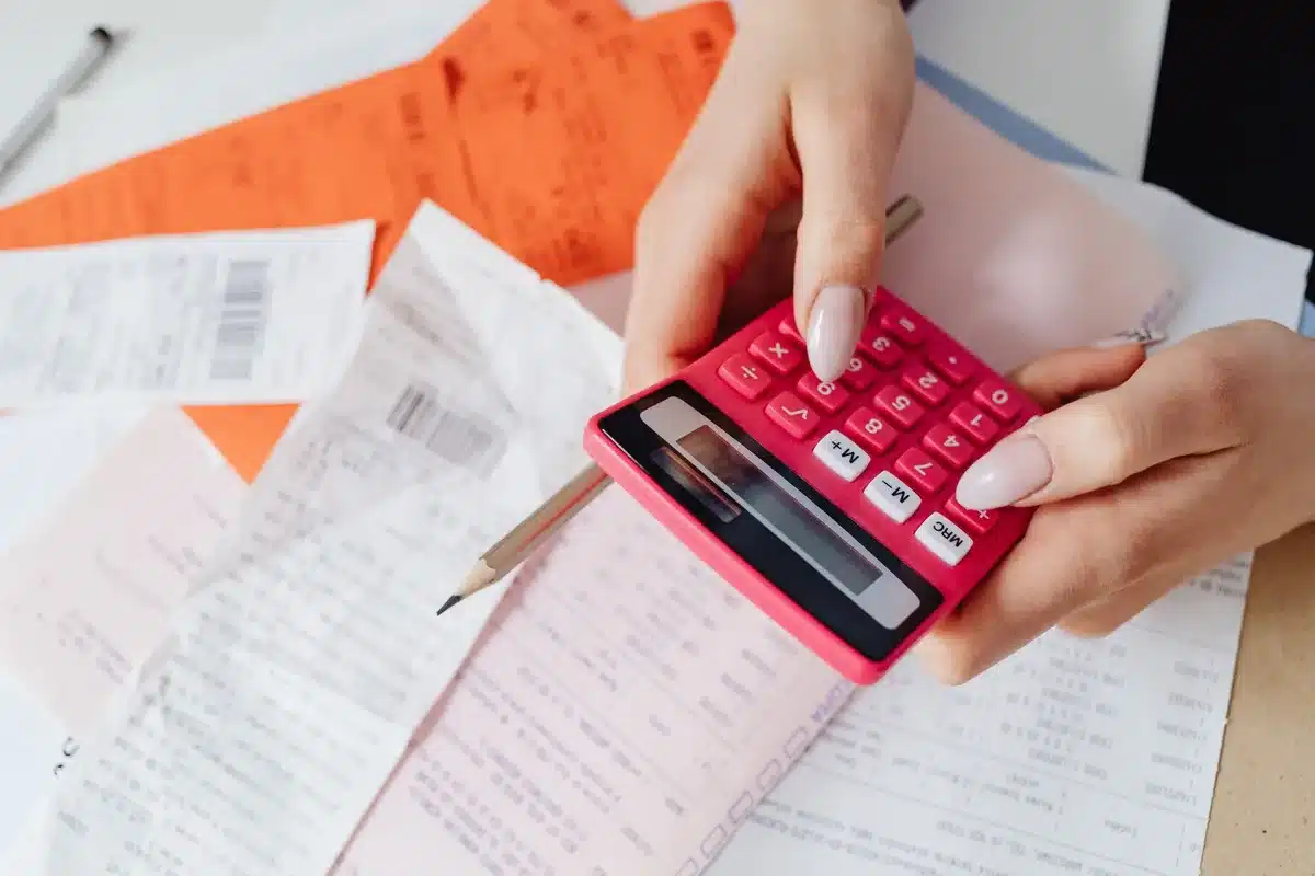Sunlit desk with rental invoice, calculator and anonymous hands checking numbers, a small stack of papers to the side